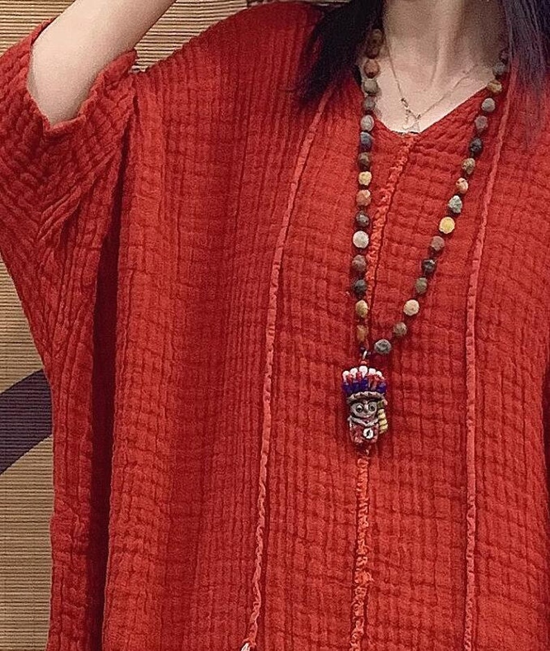May include: A red linen tunic top with a long, beaded necklace featuring a small, colorful pendant. The top has a relaxed fit and a V-neckline.