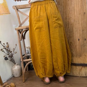 May include: Mustard yellow linen pants with two front pockets and a gathered elastic waistband. The pants have a loose, flowing design with a gathered ankle. The pants are made of a lightweight fabric.
