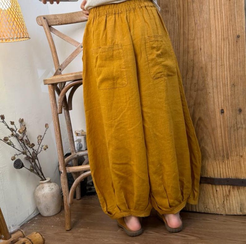 May include: Mustard yellow linen pants with two front pockets and a gathered elastic waistband. The pants have a loose, flowing design with a gathered ankle. The pants are made of a lightweight fabric.