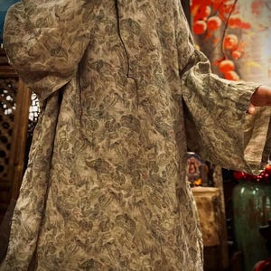 May include: A long-sleeved tunic featuring a muted olive green and grey botanical print with scattered small red accents. The garment has a loose, relaxed fit with wide sleeves, a simple round neckline, and a textured fabric appearance throughout.
