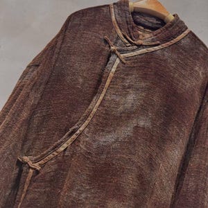 May include: A brown, long-sleeved top with a unique collar and tie closure. The top is made of a textured fabric and hangs on a wooden hanger. The design features a wrap-style front with a tie closure.