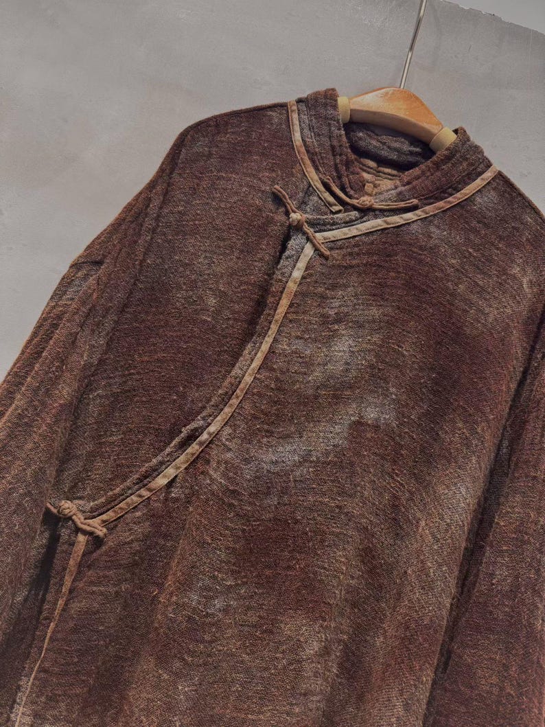 May include: A brown, long-sleeved top with a unique collar and tie closure. The top is made of a textured fabric and hangs on a wooden hanger. The design features a wrap-style front with a tie closure.