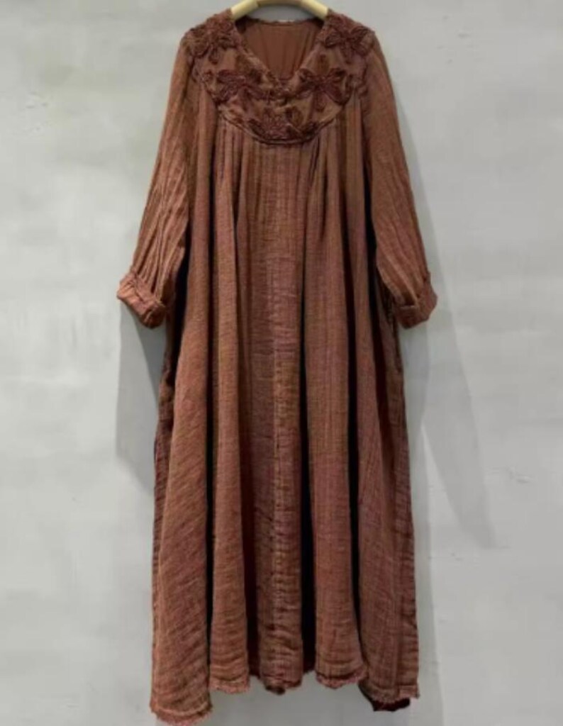 May include: A long, flowing brown dress with a V-neckline and long sleeves. The neckline features intricate floral embroidery. The dress has a loose, comfortable fit, perfect for casual wear. The fabric appears to be a lightweight, textured material.
