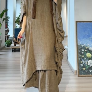 May include: A long, flowing, olive-green linen dress with a matching scarf. The dress features a loose, oversized fit with a layered skirt and gathered detailing on the sleeves. The outfit is paired with beige slip-on shoes.