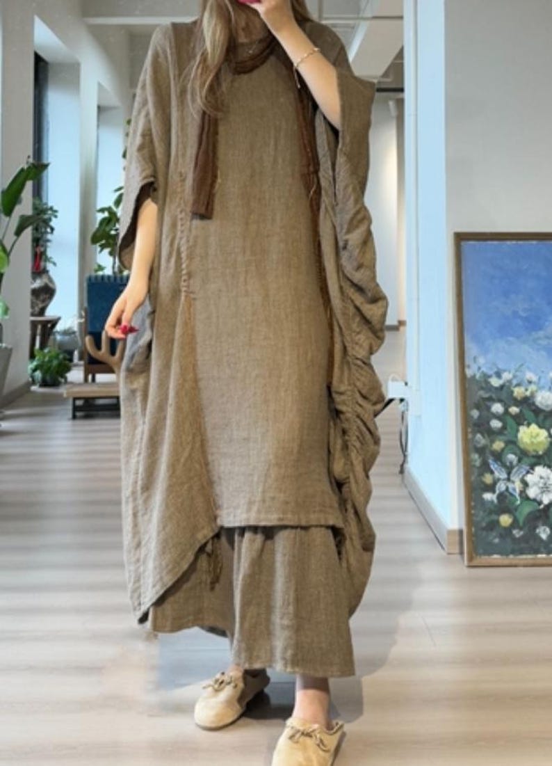 May include: A long, flowing, olive-green linen dress with a matching scarf. The dress features a loose, oversized fit with a layered skirt and gathered detailing on the sleeves. The outfit is paired with beige slip-on shoes.