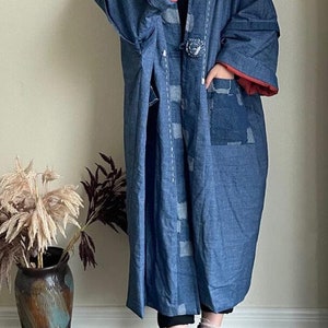Może przedstawiać: Długi, niebieski dżinsowy kimono z czerwoną podszewką i ozdobnym guzikiem. Kimono ma patchworkowy wz&oacute;r z białymi szwami i dużą kieszeń po prawej stronie.