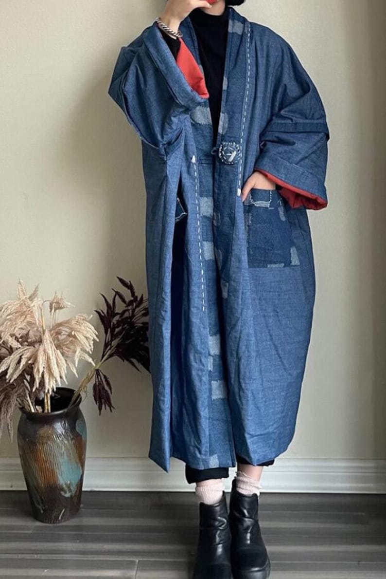 Może przedstawiać: Długi, niebieski dżinsowy kimono z czerwoną podszewką i ozdobnym guzikiem. Kimono ma patchworkowy wz&oacute;r z białymi szwami i dużą kieszeń po prawej stronie.