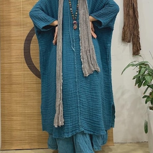 May include: A long, loose-fitting, teal blue dress with a textured fabric. The dress has long sleeves and a relaxed fit. The model is wearing a gray scarf and a beaded necklace.