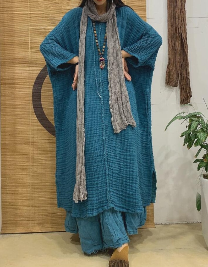May include: A long, loose-fitting, teal blue dress with a textured fabric. The dress has long sleeves and a relaxed fit. The model is wearing a gray scarf and a beaded necklace.