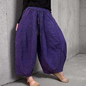 May include: Purple harem pants with a drawstring waist. The pants have a loose, baggy fit and are made of a textured fabric. The model is wearing a black long-sleeve shirt and tan shoes.