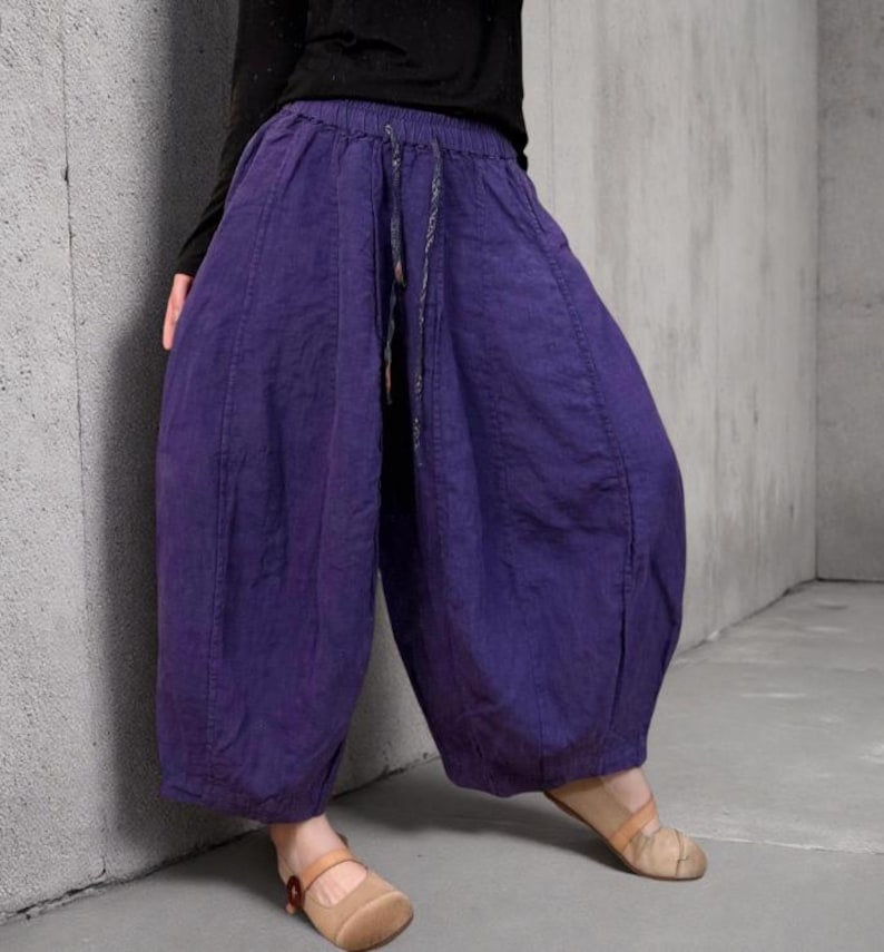 May include: Purple harem pants with a drawstring waist. The pants have a loose, baggy fit and are made of a textured fabric. The model is wearing a black long-sleeve shirt and tan shoes.