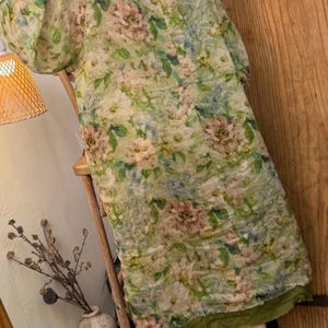 May include: A long, flowing dress with a floral pattern in shades of green, pink, and blue. The dress has a round neckline and wide sleeves. The hemline is trimmed with a green fabric. The dress is worn with brown shoes.