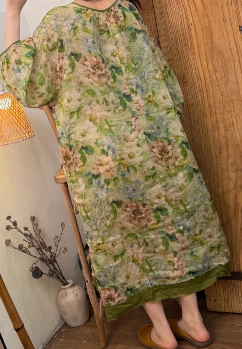 May include: A long, flowing dress with a floral pattern in shades of green, pink, and blue. The dress has a round neckline and wide sleeves. The hemline is trimmed with a green fabric. The dress is worn with brown shoes.