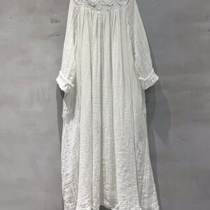 May include: A white, long-sleeved dress with a round neckline and floral embroidery. The dress has a loose, flowing silhouette and a textured fabric. The sleeves are three-quarter length with rolled cuffs. The dress falls to mid-calf length.
