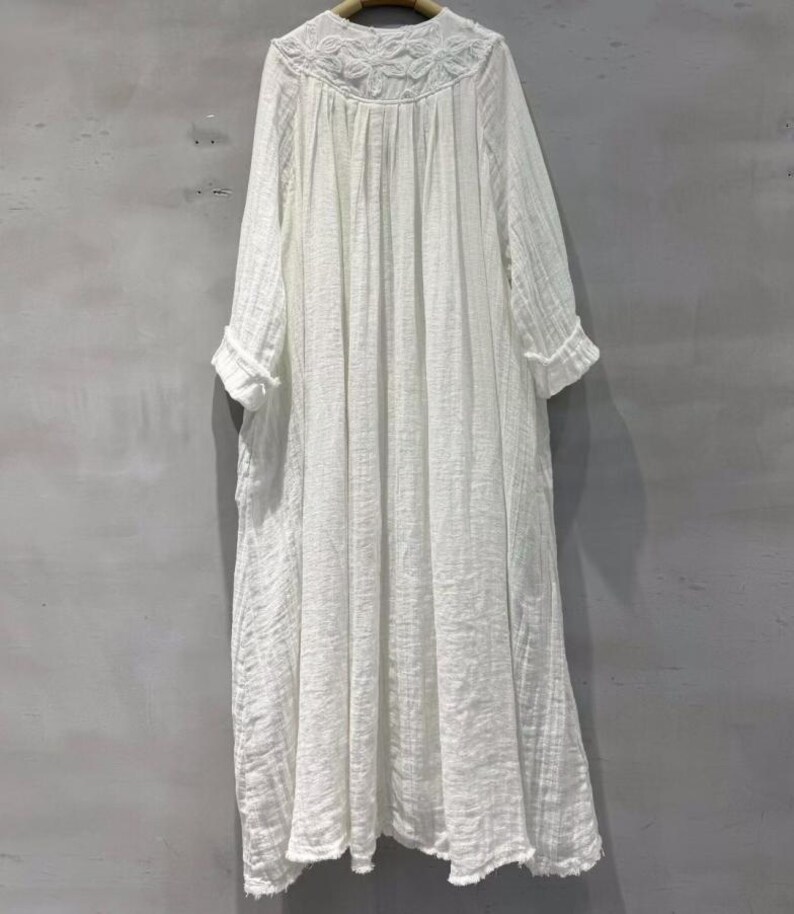 May include: A white, long-sleeved dress with a round neckline and floral embroidery. The dress has a loose, flowing silhouette and a textured fabric. The sleeves are three-quarter length with rolled cuffs. The dress falls to mid-calf length.