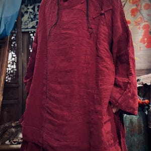 May include: A loose-fitting, long-sleeved, burgundy linen top with a mandarin collar and a button closure at the neck. The top has a relaxed fit and a slightly A-line silhouette.