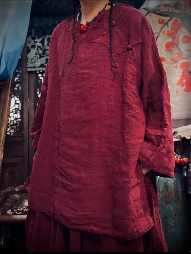 May include: A loose-fitting, long-sleeved, burgundy linen top with a mandarin collar and a button closure at the neck. The top has a relaxed fit and a slightly A-line silhouette.