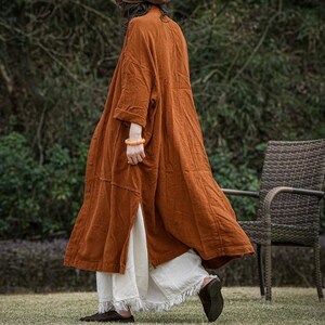 Op de afbeelding: Een lange, roestkleurige jas met een knoopsluiting aan de voorkant en zijsplitten. De jas wordt gecombineerd met een witte broek met franjes en donkere schoenen. De outfit wordt buiten gedragen, met een vage achtergrond van groen en een rieten stoel.