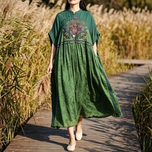 May include: A forest green midi dress featuring a high neckline with small button closures, short sleeves, and a gathered waist. The bodice displays a detailed, multi-colored floral embroidery pattern, while the skirt has a subtle textured finish.