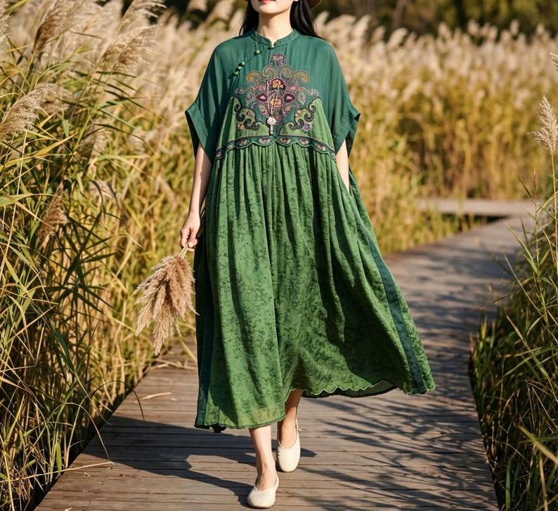 May include: A forest green midi dress featuring a high neckline with small button closures, short sleeves, and a gathered waist. The bodice displays a detailed, multi-colored floral embroidery pattern, while the skirt has a subtle textured finish.