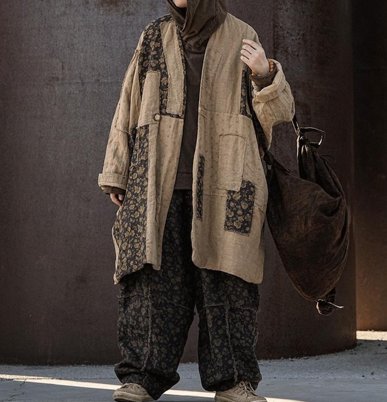 May include: A beige patchwork coat with floral accents and matching pants. The coat has a long, loose fit with dark floral patches. The pants are a dark floral pattern. A brown bag hangs from the shoulder.