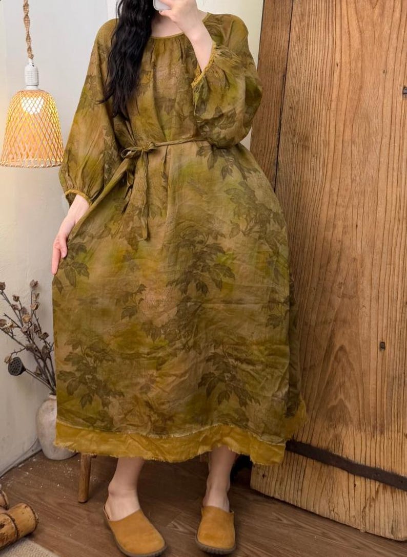 May include: A long, olive green dress with a leaf pattern. The dress has long, puffy sleeves, a round neckline, and a matching belt. The hemline and sleeve cuffs are trimmed with a mustard yellow fabric.