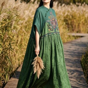 May include: A floor-length dark green dress featuring a mandarin collar with side button closures, short sleeves, and a gathered skirt. The bodice displays an intricate, multi-colored embroidered pattern in shades of purple, pink, and gold.