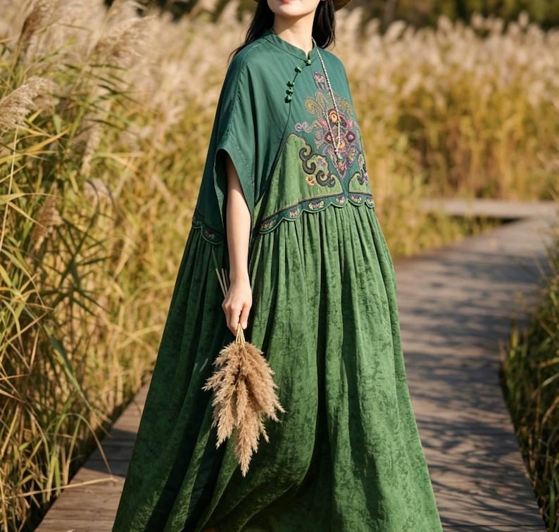 May include: A floor-length dark green dress featuring a mandarin collar with side button closures, short sleeves, and a gathered skirt. The bodice displays an intricate, multi-colored embroidered pattern in shades of purple, pink, and gold.