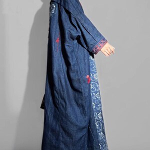 May include: A long, dark blue denim coat with embroidered details. The coat features red and white floral embroidery on the sleeves and along the front panels. The coat is floor-length and has a relaxed fit.