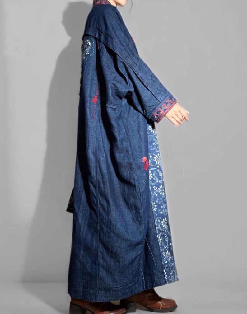 May include: A long, dark blue denim coat with embroidered details. The coat features red and white floral embroidery on the sleeves and along the front panels. The coat is floor-length and has a relaxed fit.