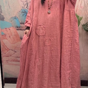 May include: A long, flowing coral dress with a relaxed fit and wide sleeves. The dress has three circular, textured patches and vertical stitching details. The fabric looks like a lightweight linen or cotton blend, ideal for warmer weather.