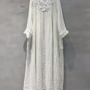 May include: A white, long-sleeved dress with a V-neckline and a textured, flowing design. The dress features intricate floral embroidery around the neckline and button details. The sleeves have ruffled cuffs, and the dress appears to be made of a lightweight fabric.