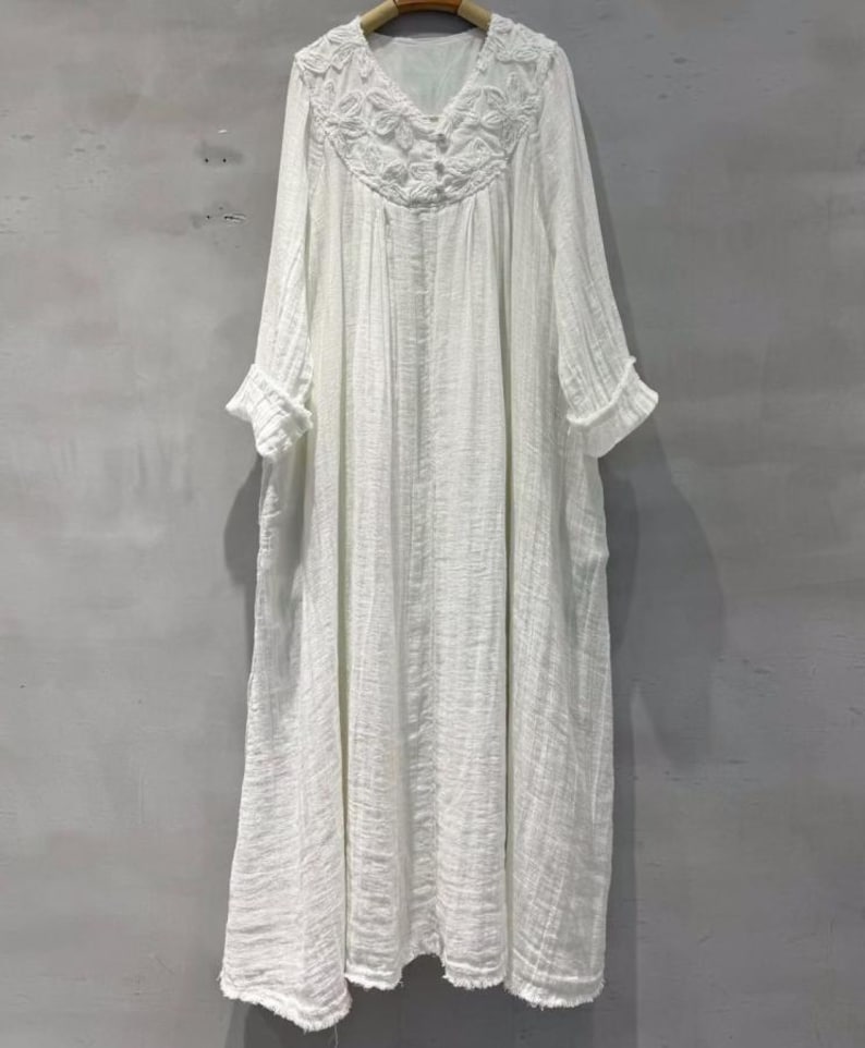 May include: A white, long-sleeved dress with a V-neckline and a textured, flowing design. The dress features intricate floral embroidery around the neckline and button details. The sleeves have ruffled cuffs, and the dress appears to be made of a lightweight fabric.