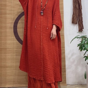 May include: A long, loose-fitting, red linen dress with a V-neckline and 3/4 sleeves. The dress has a gathered skirt and a beaded necklace with a pendant.