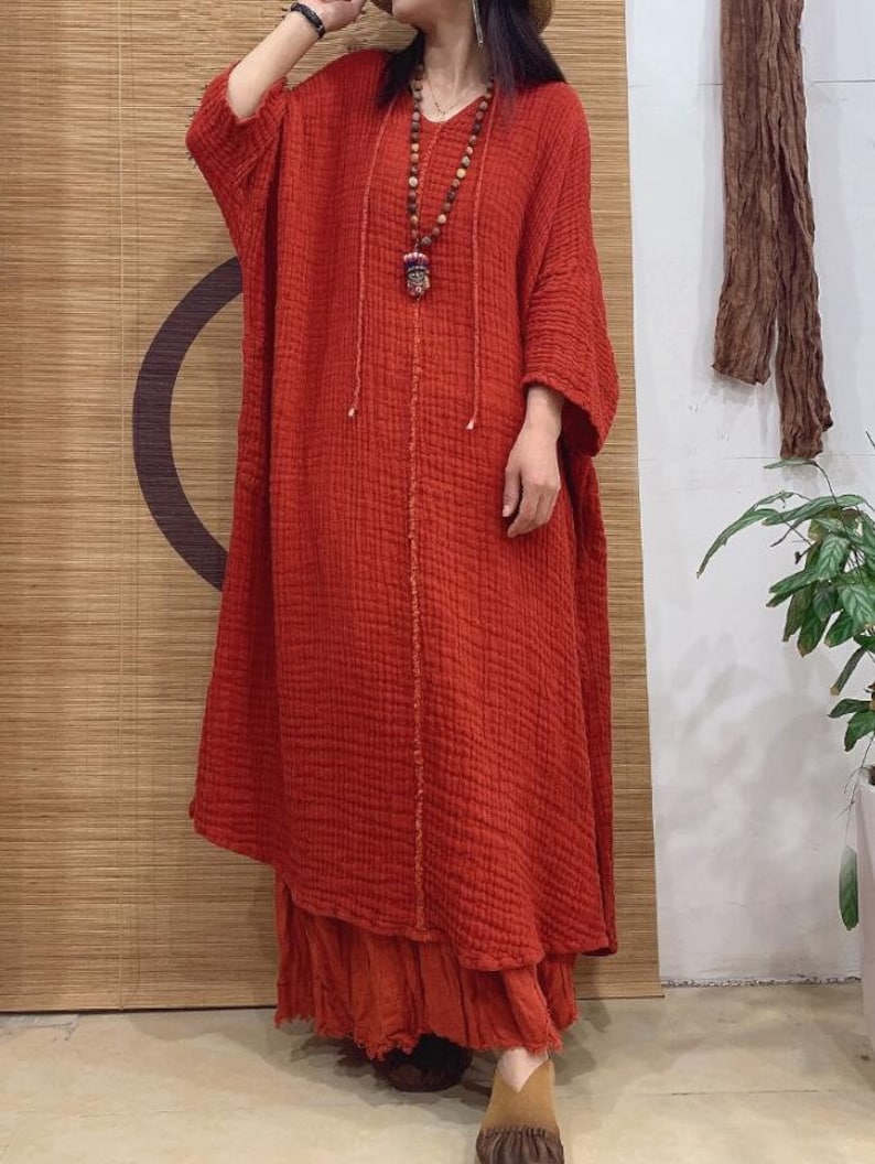 May include: A long, loose-fitting, red linen dress with a V-neckline and 3/4 sleeves. The dress has a gathered skirt and a beaded necklace with a pendant.