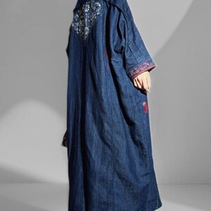 May include: A long, dark blue denim coat with intricate embroidery on the back and sleeves. The coat features a mandarin collar and a flowing, floor-length design. Red and white floral embroidery accents the cuffs and back panel, adding a touch of color.
