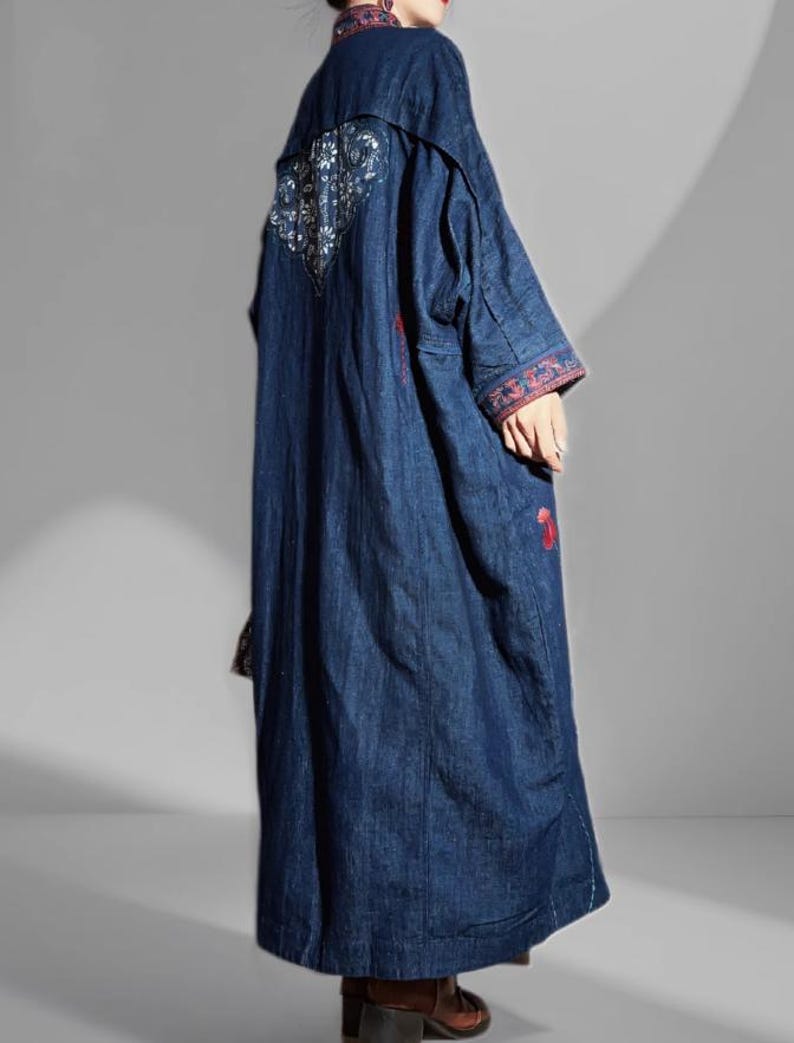 May include: A long, dark blue denim coat with intricate embroidery on the back and sleeves. The coat features a mandarin collar and a flowing, floor-length design. Red and white floral embroidery accents the cuffs and back panel, adding a touch of color.