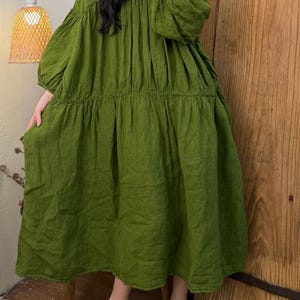 May include: A long, olive green dress with a V-neck and long, voluminous sleeves. The dress has a tiered skirt and a relaxed fit. The fabric appears to be linen or a similar lightweight material. The dress is suitable for casual wear.