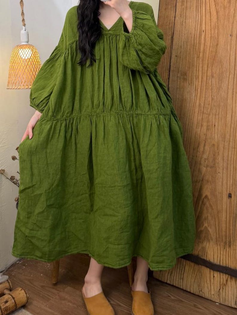 May include: A long, olive green dress with a V-neck and long, voluminous sleeves. The dress has a tiered skirt and a relaxed fit. The fabric appears to be linen or a similar lightweight material. The dress is suitable for casual wear.