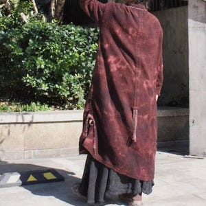 May include: A long, burgundy coat with a tie-dye effect. The coat has a textured appearance and features a unique design with pockets and a long, flowing silhouette. The coat is paired with a dark skirt.