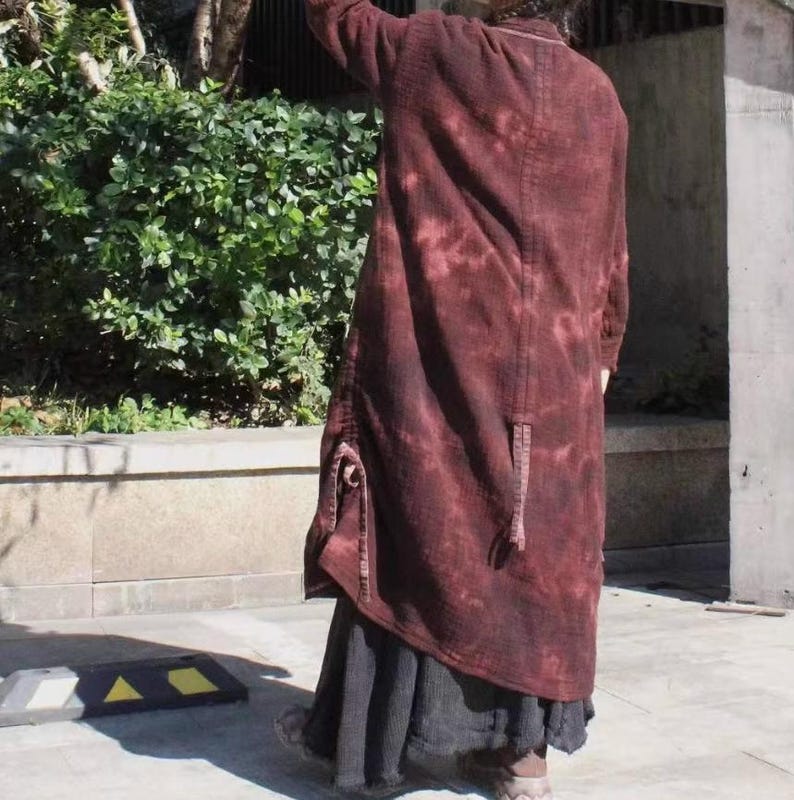 May include: A long, burgundy coat with a tie-dye effect. The coat has a textured appearance and features a unique design with pockets and a long, flowing silhouette. The coat is paired with a dark skirt.