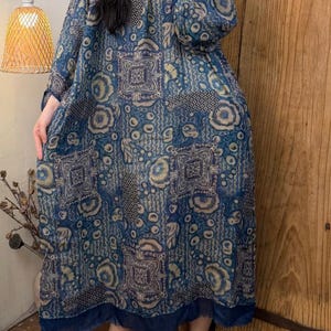 May include: A long, flowing dress in shades of blue with a repeating floral and geometric pattern. The dress has long sleeves and a round neckline. The hemline is trimmed with a darker blue fabric. The dress is made of a lightweight material.
