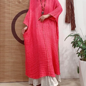 May include: A long, pink, loose-fitting dress with a V-neck and a drawstring hood. The dress is made of a textured fabric and has a relaxed fit.