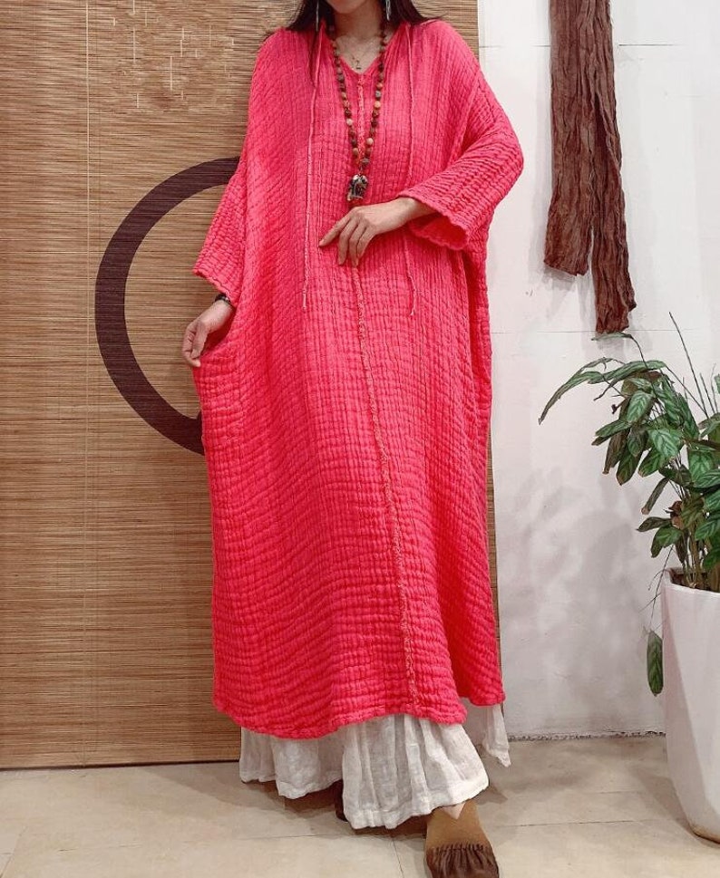 May include: A long, pink, loose-fitting dress with a V-neck and a drawstring hood. The dress is made of a textured fabric and has a relaxed fit.