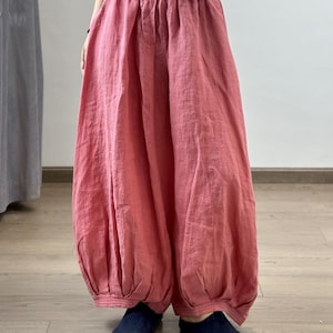 May include: Loose-fitting, coral-colored linen pants with a gathered waist and elasticized ankles. The pants have a relaxed, flowing silhouette. The fabric appears lightweight and breathable, suitable for warm weather. The pants are paired with dark blue shoes.
