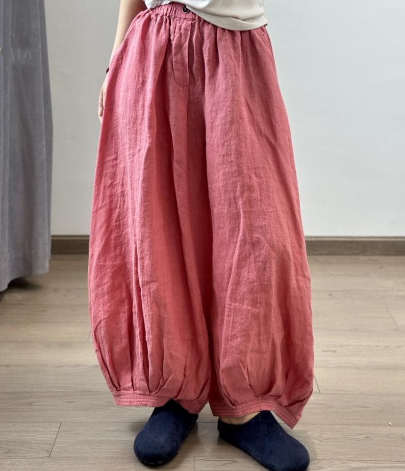May include: Loose-fitting, coral-colored linen pants with a gathered waist and elasticized ankles. The pants have a relaxed, flowing silhouette. The fabric appears lightweight and breathable, suitable for warm weather. The pants are paired with dark blue shoes.
