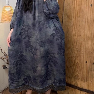 May include: A long-sleeved, loose-fitting dress with a botanical leaf pattern in shades of blue and gray. The dress has a round neckline and a flowing silhouette. The hemline falls to the ankles. The model is wearing yellow-brown suede slip-on shoes.