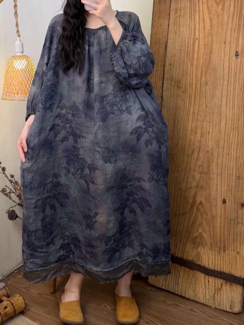 May include: A long-sleeved, loose-fitting dress with a botanical leaf pattern in shades of blue and gray. The dress has a round neckline and a flowing silhouette. The hemline falls to the ankles. The model is wearing yellow-brown suede slip-on shoes.