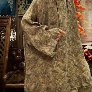May include: A loose-fitting, long-sleeved tunic featuring an all-over muted green and brown botanical leaf print. The garment has a round neckline, wide sleeves, and side slits at the hem, paired with light-colored trousers visible at the bottom.