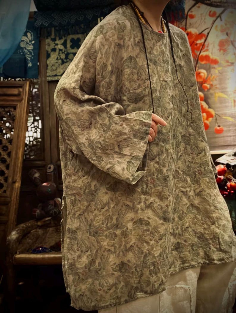 May include: A loose-fitting, long-sleeved tunic featuring an all-over muted green and brown botanical leaf print. The garment has a round neckline, wide sleeves, and side slits at the hem, paired with light-colored trousers visible at the bottom.
